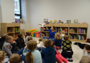 Dzieci siedzą na dywanie, pani bibliotekarka czyta dzieciom książkę.